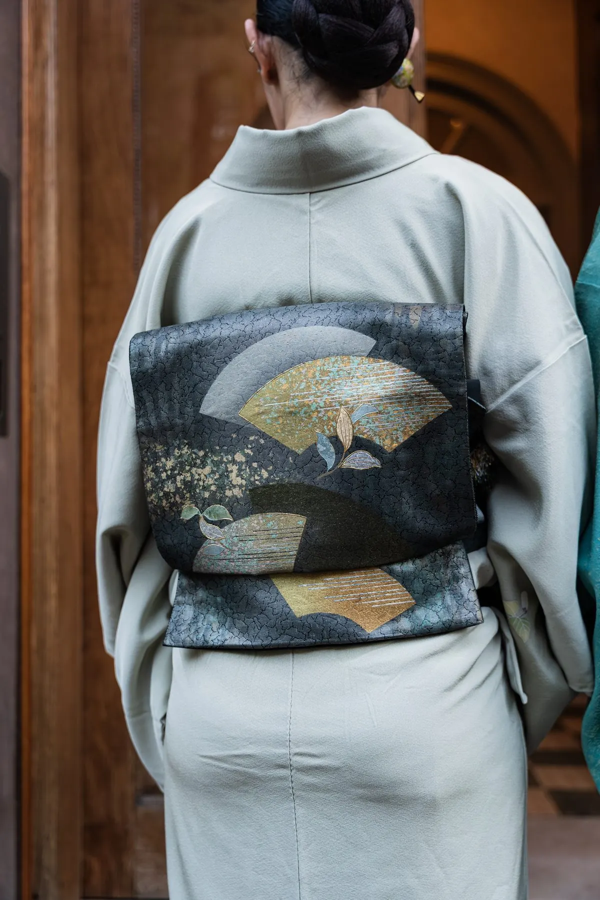 A host in pale green kimono seen from behind, a fan tucked in her obi